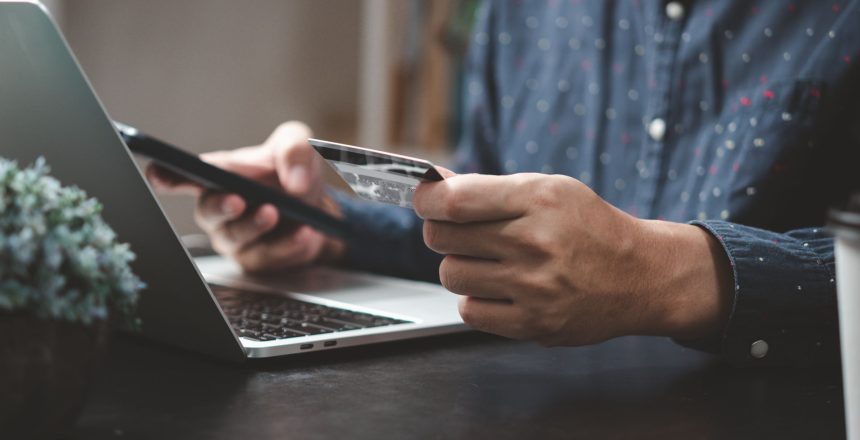 Vyriškos rankos prie nešiojamojo kompiuterio, viena ranka laiko telefoną, kita – mokėjimo kortelę.